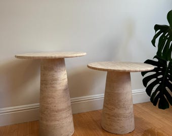 May include: Two beige stone side tables with round tops and conical bases. The tables are made of a light-colored stone with natural veining. The tables are of different sizes and are placed on a wooden floor.