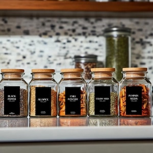 Peut inclure: Six bocaux en verre pour épices avec des couvercles en liège et des étiquettes noires. Les étiquettes sont en texte blanc et comprennent les noms "Poivre noir", "Piment", "Poudre de chili", "Graines de céleri" et "Graines de citrouille".