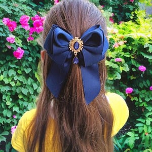Puede incluir: Un lazo para el cabello azul marino con un centro de filigrana dorado y un colgante de lágrima azul. El lazo se coloca sobre cabello castaño y una camisa amarilla, con un fondo de follaje verde y flores rosas.