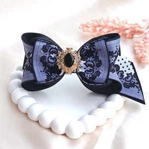 Peut inclure: Un nœud décoratif pour les cheveux avec un motif floral noir et gris et un bijou doré au centre. Le nœud est posé sur une base blanche perlée en forme de collier, sur un fond blanc doux.
