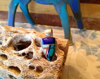 May include: A silver pendant with a geometric design featuring turquoise, lapis lazuli, and opal stones. The pendant is in the shape of a shield and has a silver bail.
