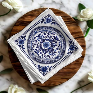 May include: A pile of white square napkins with a blue floral pattern, presented on a round wooden tray. The napkins display a central floral design and border details. White roses are placed around the tray.