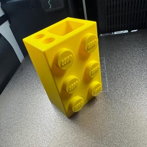 May include: A bright yellow pen holder shaped like a large building block. The block has raised circular studs with the word "LEGO" embossed on them. The holder has two circular holes and a rectangular opening.