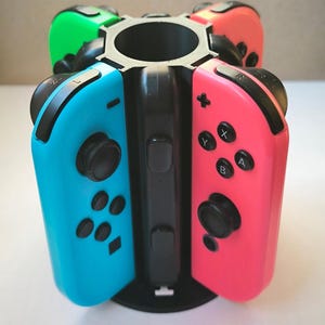 May include: A black plastic stand with four slots for holding Nintendo Switch Joy-Con controllers. The stand has a circular base and a cylindrical centre. The controllers are in different colours: blue, red, green, and orange.