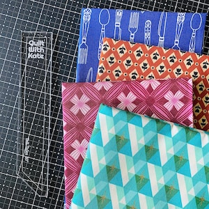 May include: Four folded fabric squares with distinct patterns: blue with white cutlery, orange with black tulips, magenta with pink geometric shapes, and teal with white and green diamonds. A clear acrylic ruler labeled Quilt With Kate 10 inches.