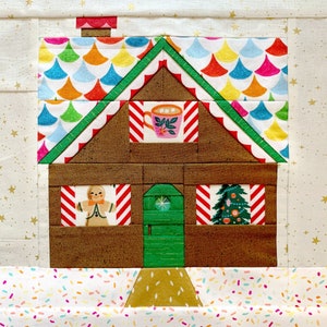May include: A gingerbread house quilt block with a green door, candy cane windows, and a Christmas tree. The roof is made of colorful fabric with a scalloped edge. The house is sitting on a white background with sprinkles.