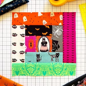 May include: A square fabric quilt block featuring a patchwork of Halloween-themed prints including ghosts, bats, skeletons, vampire teeth, and a central illustration of a cat in a dress, surrounded by various fabric scraps and a yellow rotary cutter.