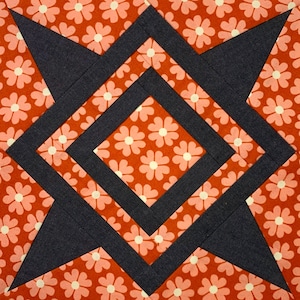 Pode incluir: Um bloco de colcha com um desenho de estrela no centro. A estrela é feita de tecido azul escuro e é cercada por um tecido vermelho com um padrão floral branco. O bloco é feito de várias camadas de tecido.