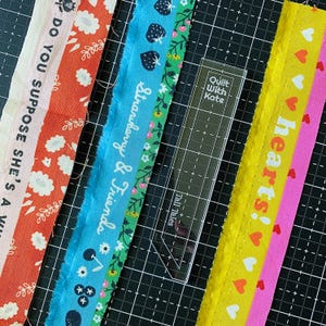 May include: Three strips of patterned fabric and a clear acrylic quilting ruler. The left strip is orange with white flowers and text. The middle strip is blue with strawberry graphics and text. The right strip is yellow with red and white hearts.