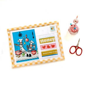 Op de afbeelding: Een gequilt kunstwerk met een blauw-witte illustratie van koks die koken, en het woord "Honey" met aardbeiengraphics. Een kleine rode schaar en een decoratieve speld zijn ook aanwezig.