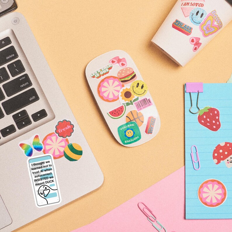 May include: A collection of colorful stickers with various designs, including a smiley face, a watermelon slice, and a burger. The stickers are arranged on a white mouse, a laptop, and a notepad. The text on the stickers includes "I AM LOVED" and "YOU ARE ENOUGH".
