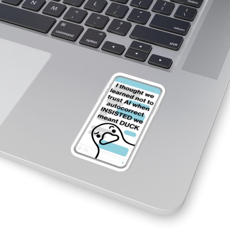 May include: A white sticker featuring a cartoon duck and text that reads "I thought we learned not to trust AI when autocorrect INSISTED we meant DUCK." The sticker is placed on a laptop.