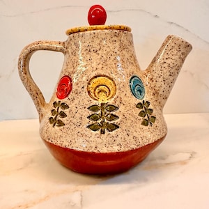 May include: A ceramic teapot with a speckled cream glaze, featuring three embossed flower motifs in red, yellow, and blue with green stems. The base is solid red, and the lid has a bright red spherical knob handle on top of a speckled lid.