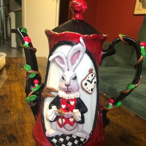 Könnte beinhalten: Eine rote und schwarze Teekanne mit einer weißen Hasenfigur auf der Vorderseite. Der Hase trägt eine Weste und hält eine Taschenuhr. Die Teekanne hat einen rankenartigen Griff mit grünen Blättern und roten Beeren.