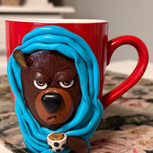 Könnte beinhalten: Eine rote Keramiktasse mit einem braunen Bären, der einen blauen Schal trägt und eine kleine Tasse hält. Der Bär hat einen mürrischen Gesichtsausdruck.