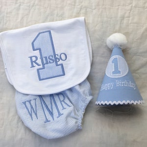 May include: A baby's first birthday set, including a bib, bloomers, and party hat. The bib and bloomers are blue and white striped, with embroidered initials and the number 1. The party hat is light blue with a white pom-pom and the words "Happy Birthday".