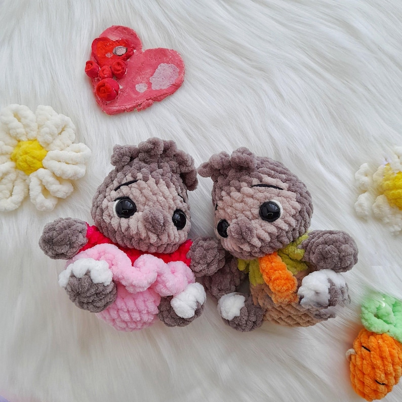 Op de afbeelding: Twee handgemaakte gehaakte knuffels met grote zwarte ogen, &eacute;&eacute;n met een roze en rode outfit, de andere met een groene en oranje sjaal. Een rood hart en twee witte madeliefachtige bloemen zijn ook zichtbaar.