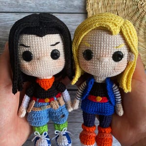 May include: Two handmade crochet dolls featuring black and yellow yarn hair respectively. The left doll wears a black shirt, orange scarf, and blue shorts. The right doll wears a black shirt, blue vest, and blue skirt with orange shoes.