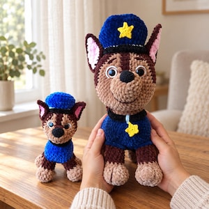 Puede incluir: Dos peluches tejidos a mano que representan a un perro marrón y beige con gorra y uniforme de policía azul. El peluche más grande tiene un emblema de estrella amarilla en la gorra y el collar, mientras que el más pequeño tiene una gorra azul.