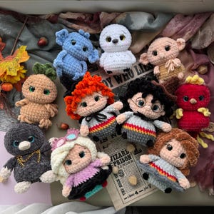 May include: A collection of ten small, handmade crochet plush dolls featuring various characters with distinct hair colors, outfits, and accessories. The dolls are crafted from soft, textured yarn and include figures with glasses, capes, and colorful clothing items arranged on a printed paper background.