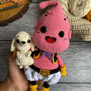 May include: Handmade crochet doll of a pink character with black eyes, yellow boots, and a purple scarf, holding a small white crochet dog. The doll has a black belt with a gold "M" on it. The background includes yarn and crochet tools.
