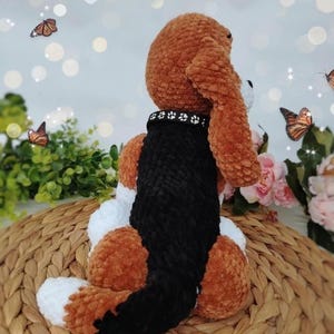 Peut inclure: Un jouet en peluche fait &agrave; la main en marron, noir et blanc, avec un collier noir orn&eacute; d'empreintes de pattes. Le chien est assis sur une surface tiss&eacute;e, avec des papillons et des fleurs en arri&egrave;re-plan.