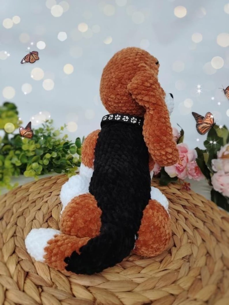 Peut inclure: Un jouet en peluche fait &agrave; la main en marron, noir et blanc, avec un collier noir orn&eacute; d'empreintes de pattes. Le chien est assis sur une surface tiss&eacute;e, avec des papillons et des fleurs en arri&egrave;re-plan.
