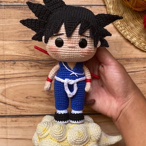 Könnte beinhalten: Handgefertigte Häkelpuppe einer Figur mit stacheligen schwarzen Haaren, die einen blauen Overall mit weißem Gürtel und schwarzen Schuhen trägt. Die Puppe steht auf einer gelben Wolke. Die Puppe hat schwarze Augen und rote Armbänder.