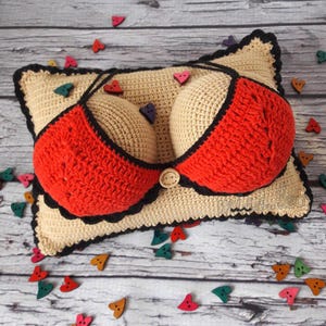 Könnte beinhalten: Ein handgefertigtes Häkelkissen mit beigefarbener Basis und einem rot-schwarzen Bikini-Oberteil-Design. Das Kissen hat zwei beige gehäkelte Brustformen und ist mit herzförmigen Knöpfen in verschiedenen Farben verziert. Das Kissen liegt auf einer Holzoberfläche.