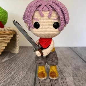 Puede incluir: Muñeco de ganchillo hecho a mano de un personaje con pelo morado, ojos negros y una bufanda roja, sosteniendo una espada plateada. El muñeco lleva pantalones cortos marrones y botas amarillas.