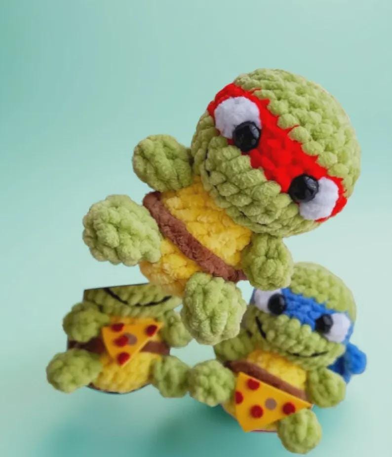 May include: Crocheted Teenage Mutant Ninja Turtles in green, yellow, and brown. Each turtle has a different colored mask: red, blue, and brown. They are holding pizza slices. The turtles are handmade.