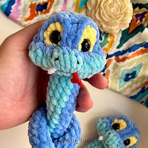 Op de afbeelding: Een handgemaakt blauw en turquoise gehaakt slangenknuffel met gele ogen en een rode tong. De pluche slang is gemaakt van zacht, getextureerd garen. De slang is een grillig speelgoed.