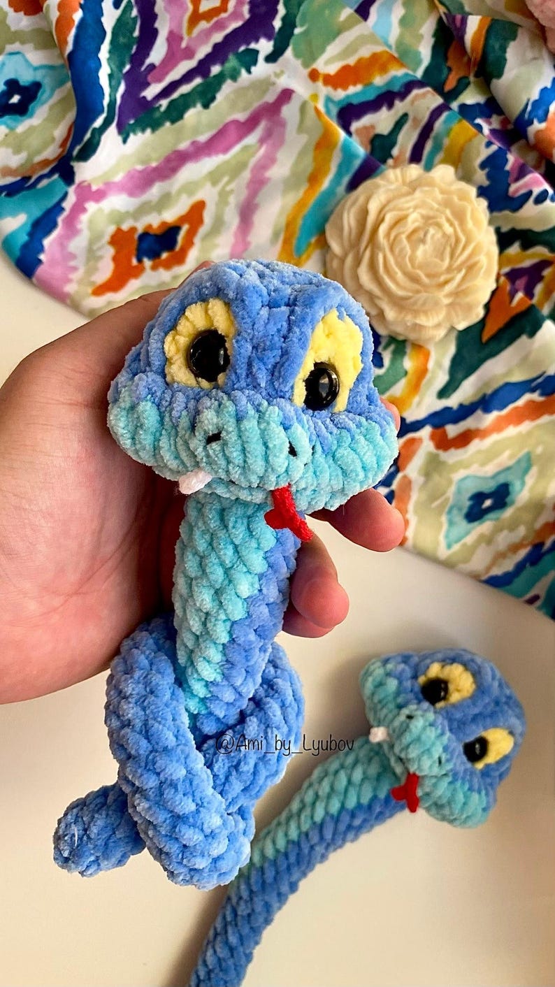 Op de afbeelding: Een handgemaakt blauw en turquoise gehaakt slangenknuffel met gele ogen en een rode tong. De pluche slang is gemaakt van zacht, getextureerd garen. De slang is een grillig speelgoed.