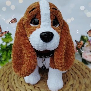 Peut inclure: Un chien en peluche fait &agrave; la main, avec une fourrure marron et blanche, de grandes oreilles tombantes et un nez noir. Le chien porte un collier noir avec une petite cloche. La peluche est pos&eacute;e sur une surface tiss&eacute;e, avec des papillons en arri&egrave;re-plan.