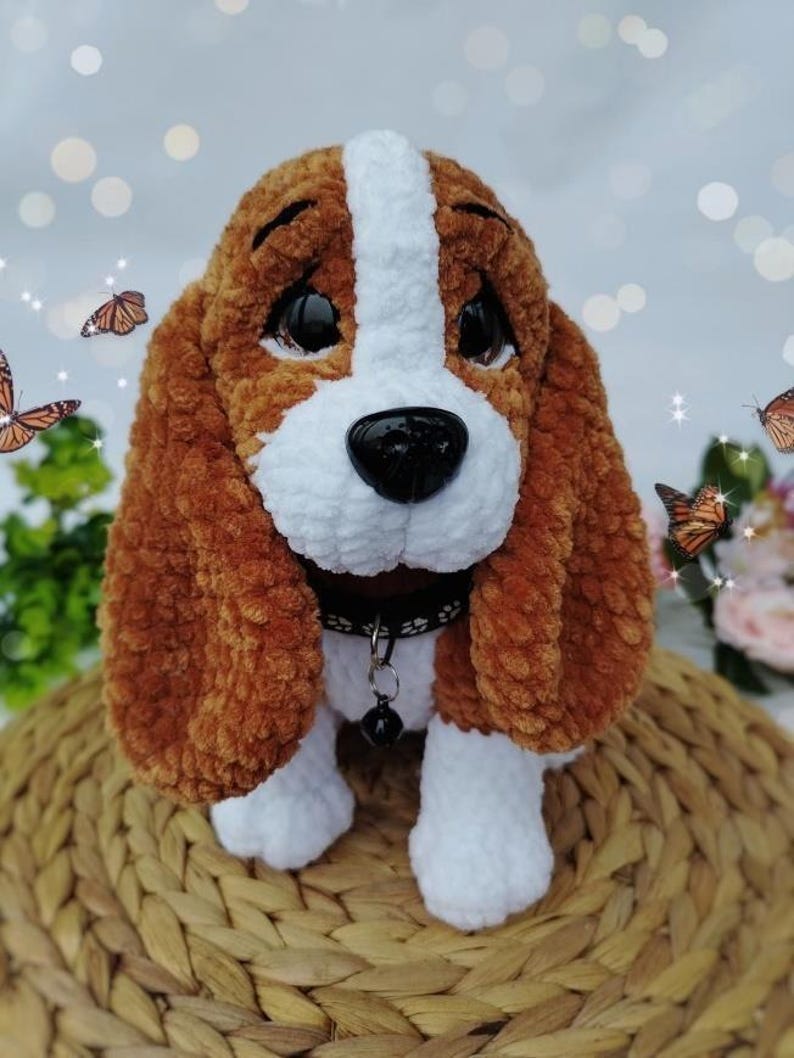 Peut inclure: Un chien en peluche fait &agrave; la main, avec une fourrure marron et blanche, de grandes oreilles tombantes et un nez noir. Le chien porte un collier noir avec une petite cloche. La peluche est pos&eacute;e sur une surface tiss&eacute;e, avec des papillons en arri&egrave;re-plan.