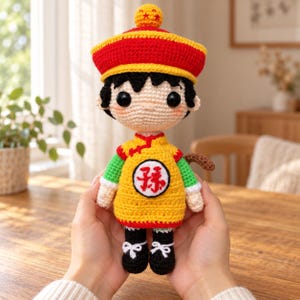 Puede incluir: Una muñeca de ganchillo que representa un personaje con pelo negro, vistiendo una túnica amarilla con mangas verdes, un sombrero rojo y amarillo con una pequeña bola en la parte superior y zapatos negros. La túnica tiene un parche circular.