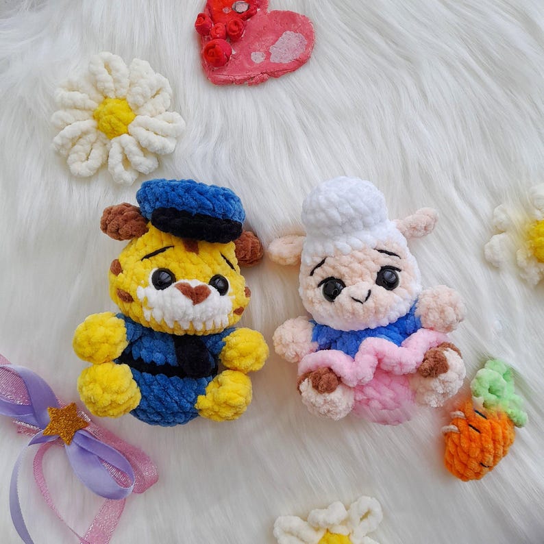 Op de afbeelding: Handgemaakte gehaakte speelgoed, waaronder een gele tijger met een blauwe matrozenhoed en -uniform, en een wit schaap met een koksmuts en roze rok. Extra gehaakte bloemen, een hart en een wortel zijn ook zichtbaar.