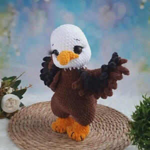 May include: A crocheted bald eagle plush toy with a white head, brown body, and orange beak and feet. The eagle has large dark eyes with long black eyelashes and wings spread outward. The texture of the yarn is soft and plush throughout.