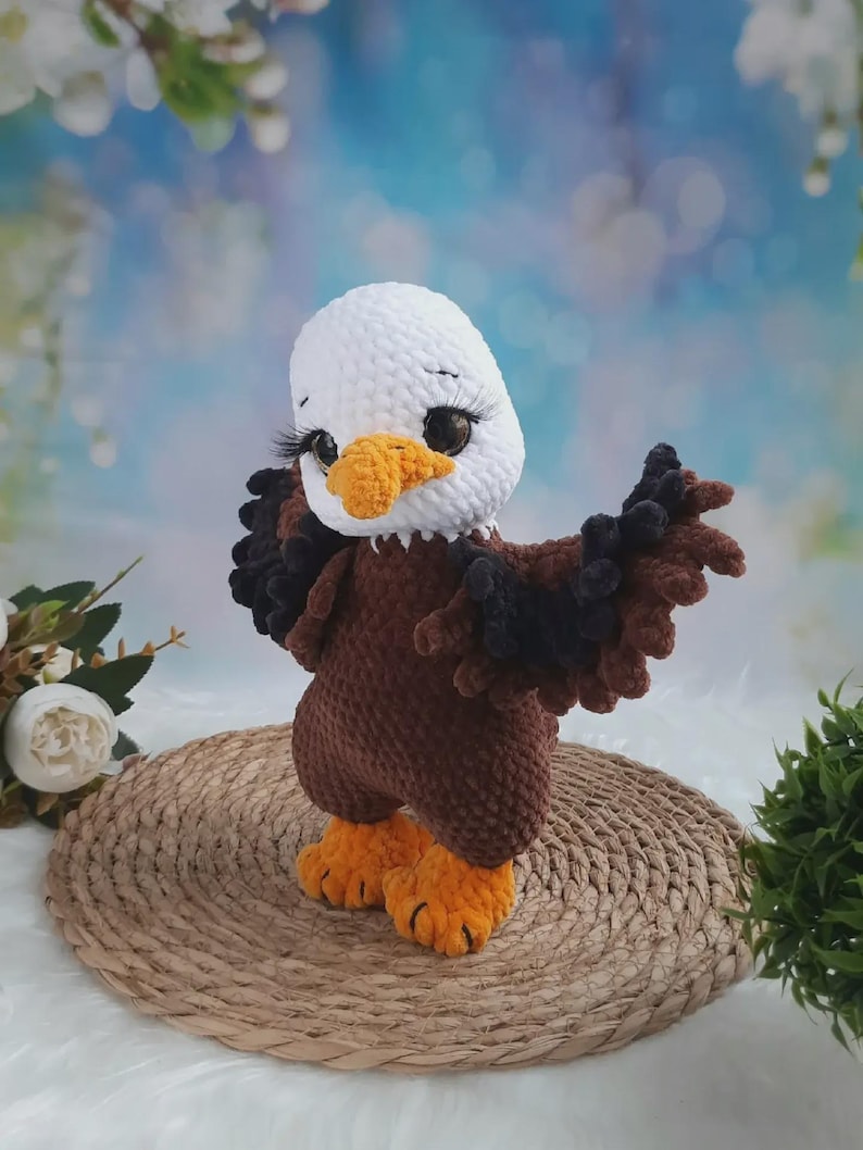 May include: A crocheted bald eagle plush toy with a white head, brown body, and orange beak and feet. The eagle has large dark eyes with long black eyelashes and wings spread outward. The texture of the yarn is soft and plush throughout.