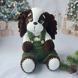 May include: A crocheted dog toy with a white and brown head, brown ears, and green dungarees. The dog has a white chest and paws, and a small brown bow tie. The toy is positioned in front of a Christmas tree.