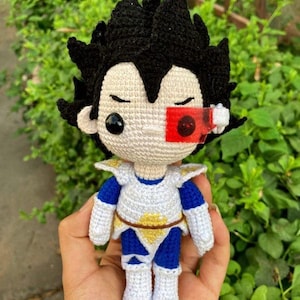 Puede incluir: Muñeco de ganchillo de un personaje con pelo negro puntiagudo, ojos cerrados y una visera roja. El muñeco lleva un atuendo blanco y azul con detalles amarillos. El muñeco mide aproximadamente 13 cm de alto.