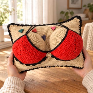 May include: A rectangular beige crochet pillow featuring two rounded, stuffed protrusions covered by a red crochet bikini top with black trim. The design includes small, colorful heart-shaped buttons and a central wooden button with text.
