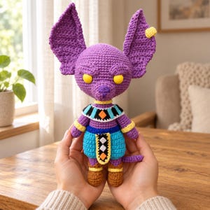 Könnte beinhalten: Eine violette Häkelpuppe mit großen spitzen Ohren, gelben Augen und einem Kragen mit blauen, schwarzen und orangefarbenen geometrischen Mustern. Die Puppe trägt einen blauen Rock mit einem zentralen schwarz-orangen Tabard und braune Schuhe.
