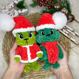 Puede incluir: Dos peluches verdes de personajes navideños con gorros rojos de Papá Noel y pompones blancos. Uno lleva una chaqueta roja, el otro una bufanda roja. Los juguetes tienen expresiones enfadadas y se sostienen sobre una esterilla tejida.
