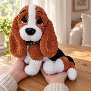 Peut inclure: Une peluche en forme de chien basset, fabriquée en fil doux marron, blanc et noir. Le jouet présente de longues oreilles tombantes, de grands yeux sombres, un nez noir et un collier noir orné d'un motif d'empreintes de pattes blanches.
