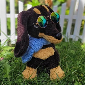 Puede incluir: Un perro salchicha de ganchillo hecho a mano con pelaje negro y canela, con gafas de sol redondas y una bufanda azul. El perro está posando sobre hierba verde frente a una valla blanca, creando una escena caprichosa.