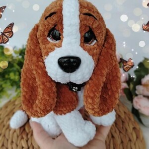 Peut inclure: Un chien en peluche fait &agrave; la main, avec une fourrure marron et blanche, de grands yeux noirs et un nez noir. Le chien a des oreilles tombantes et un petit collier noir. La peluche est pos&eacute;e sur une surface tiss&eacute;e, avec des papillons en arri&egrave;re-plan.