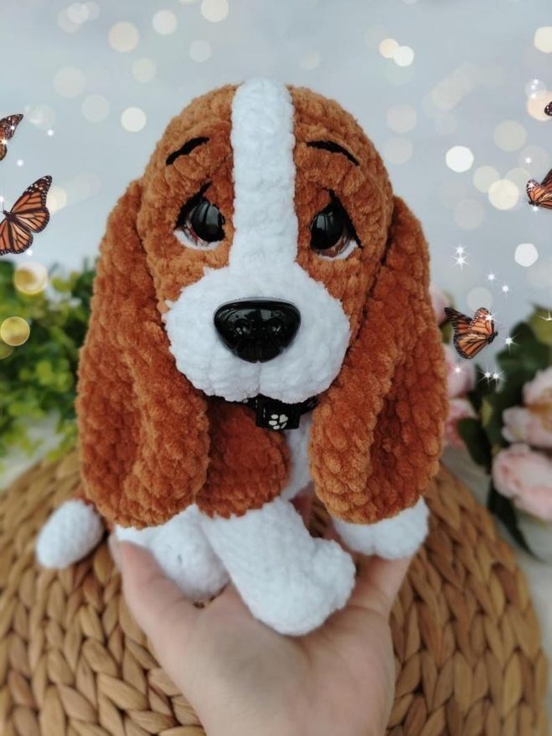 Peut inclure: Un chien en peluche fait &agrave; la main, avec une fourrure marron et blanche, de grands yeux noirs et un nez noir. Le chien a des oreilles tombantes et un petit collier noir. La peluche est pos&eacute;e sur une surface tiss&eacute;e, avec des papillons en arri&egrave;re-plan.