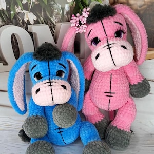 May include: Two plush donkey toys, one blue and one pink, are displayed. The blue donkey has grey accents and the pink donkey has a flower accessory. Both have large ears, sad eyes, and grey pom-pom feet.