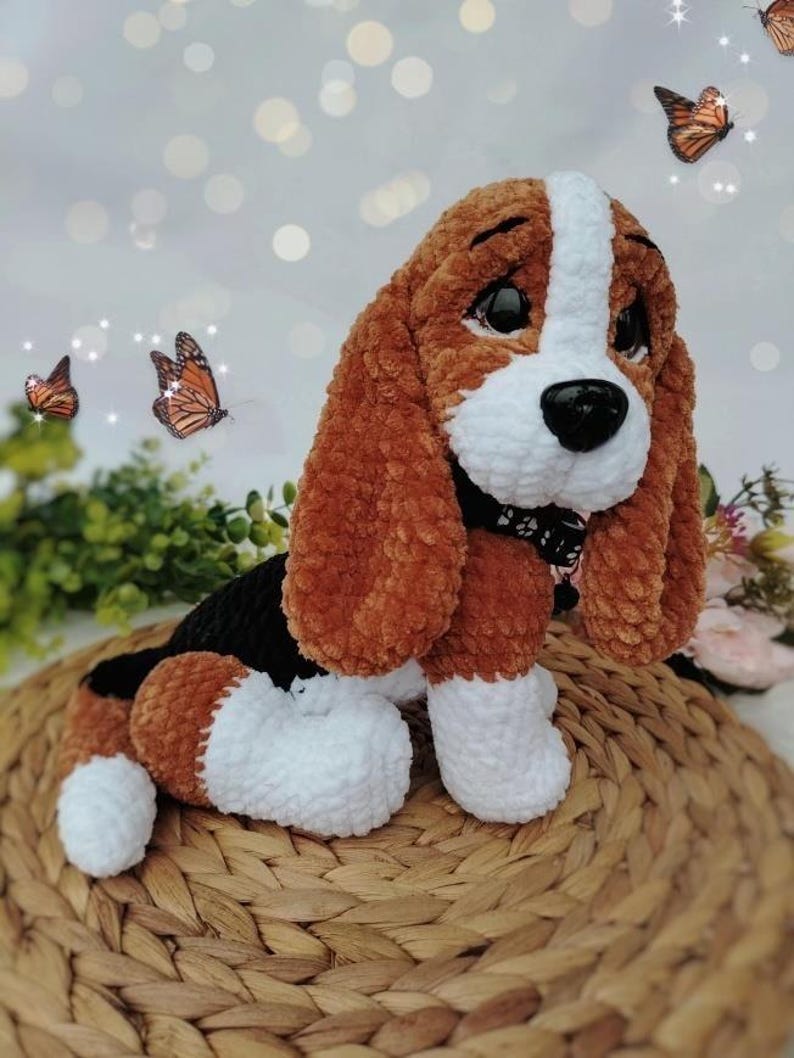 Peut inclure: Un chien en peluche fait &agrave; la main, avec des accents marron, blancs et noirs. Le chien a de grandes oreilles tombantes, un museau blanc et un nez noir. Il est assis sur un tapis tress&eacute;, avec des papillons en arri&egrave;re-plan. Le chien mesure environ 25 cm de haut.